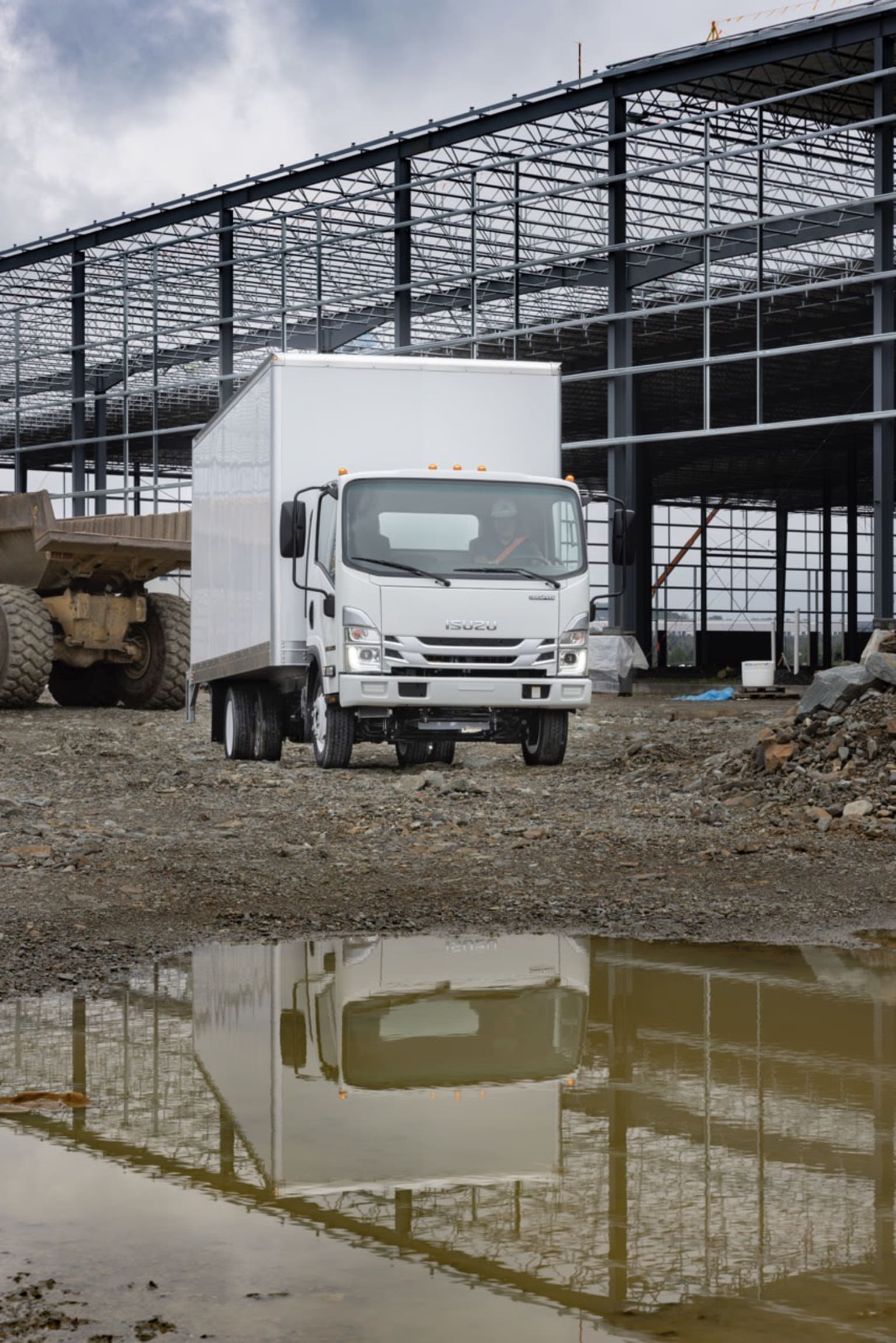 A white delivery truck is parked in the foreground, reflected in a puddle on the ground. In the background, a large metal-framed structure, likely a warehouse or industrial building, stands against a cloudy sky.