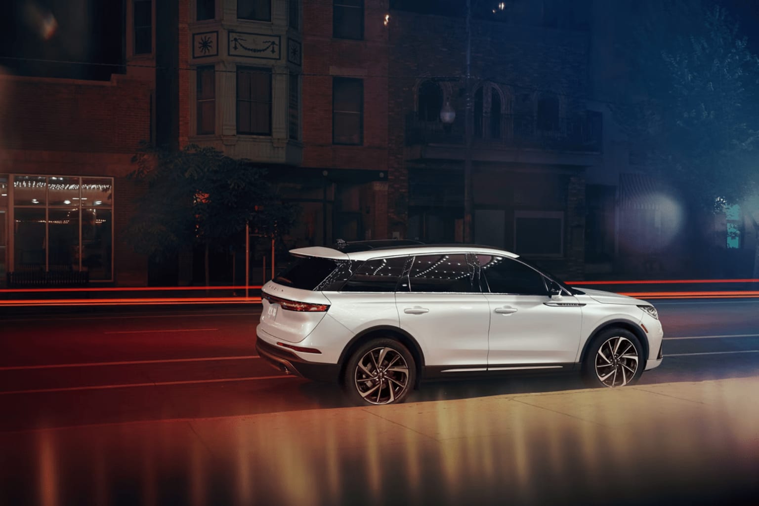 White SUV parked on city street at night with blurred lights and buildings in the background.
