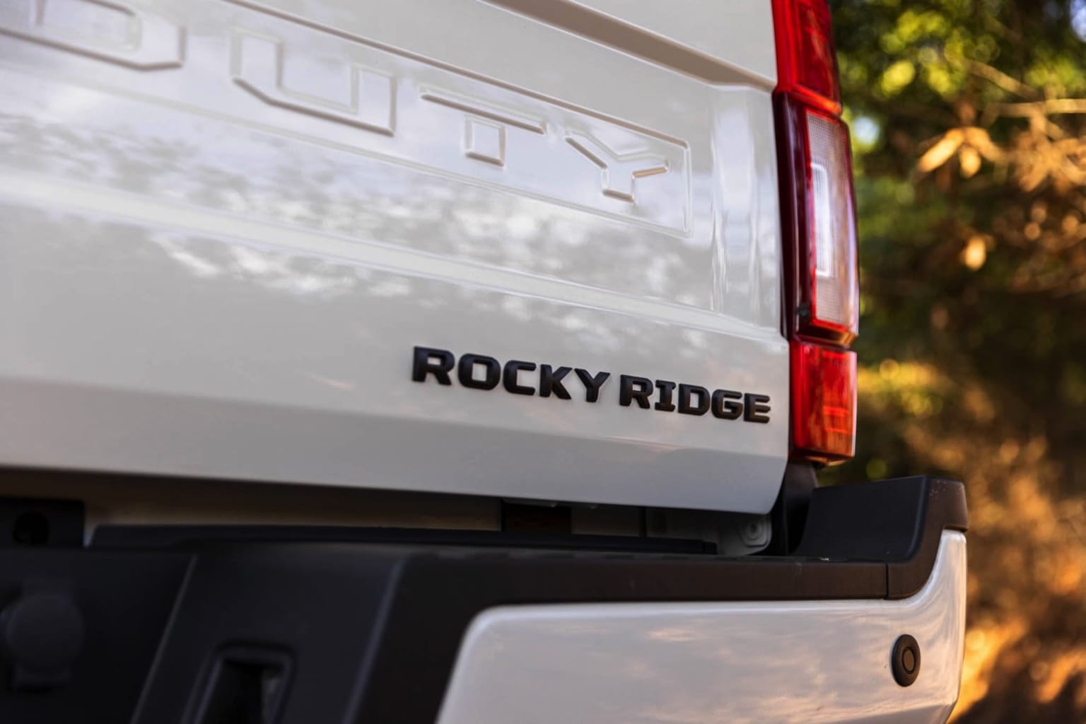 The image shows the rear of a white recreational vehicle set against a blurred natural background with trees and sunlight.