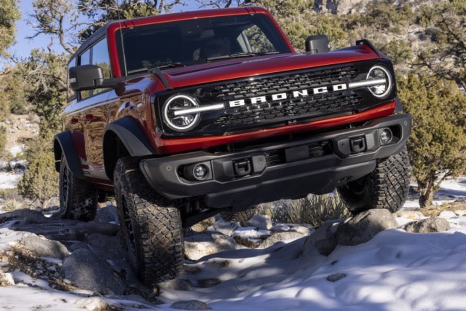 A red SUV is shown navigating through a snowy, rocky terrain with trees and bushes in the background.