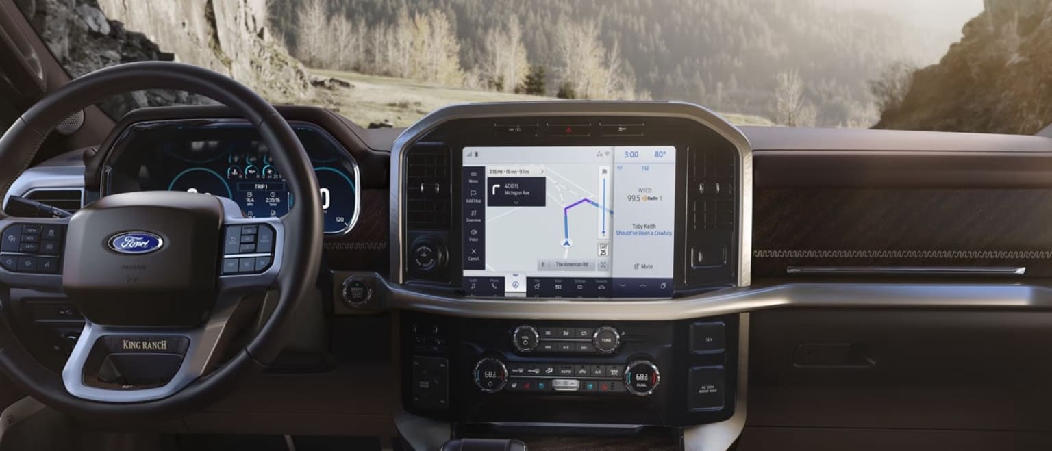 Interior of a vehicle with dashboard display and controls, with backdrop of mountainous landscape.