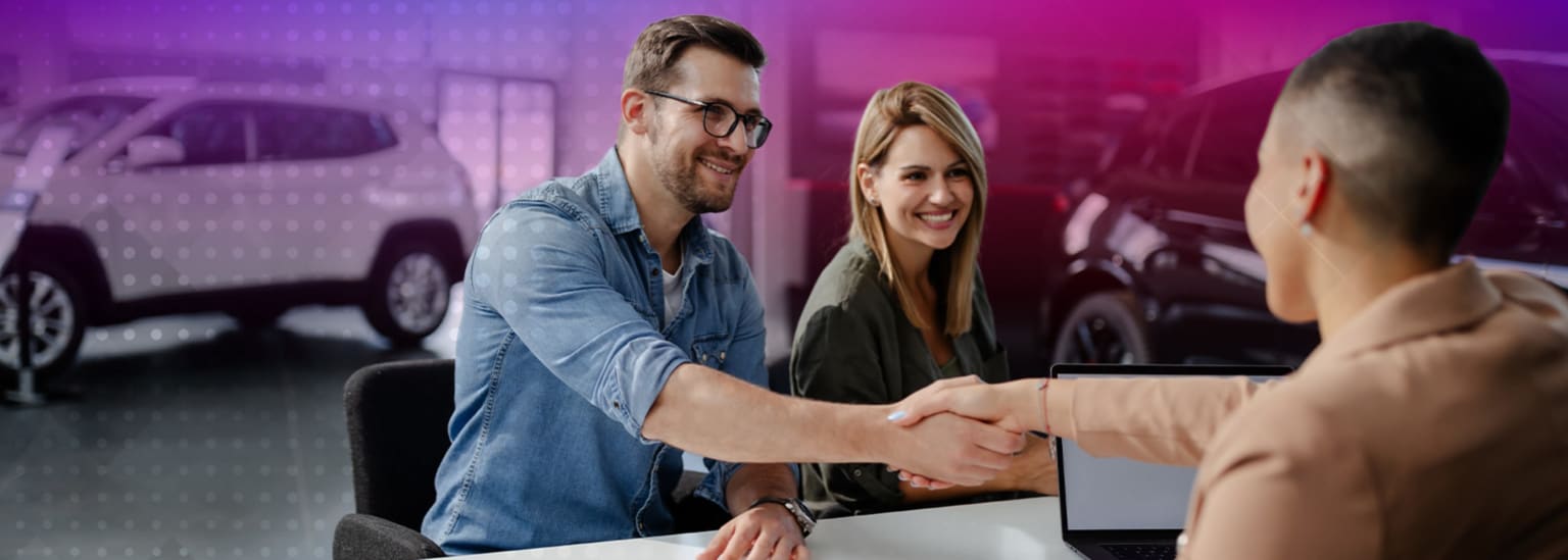 Two people shaking hands in an office with a car in the background.
