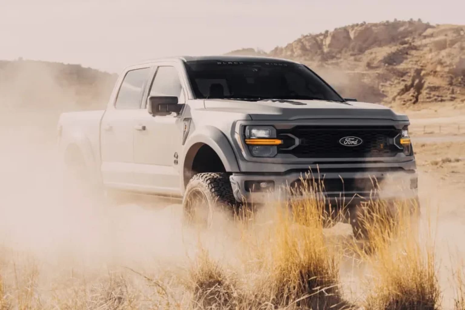 A white pickup truck kicks up dust in the foreground against a backdrop of arid, rocky hills.