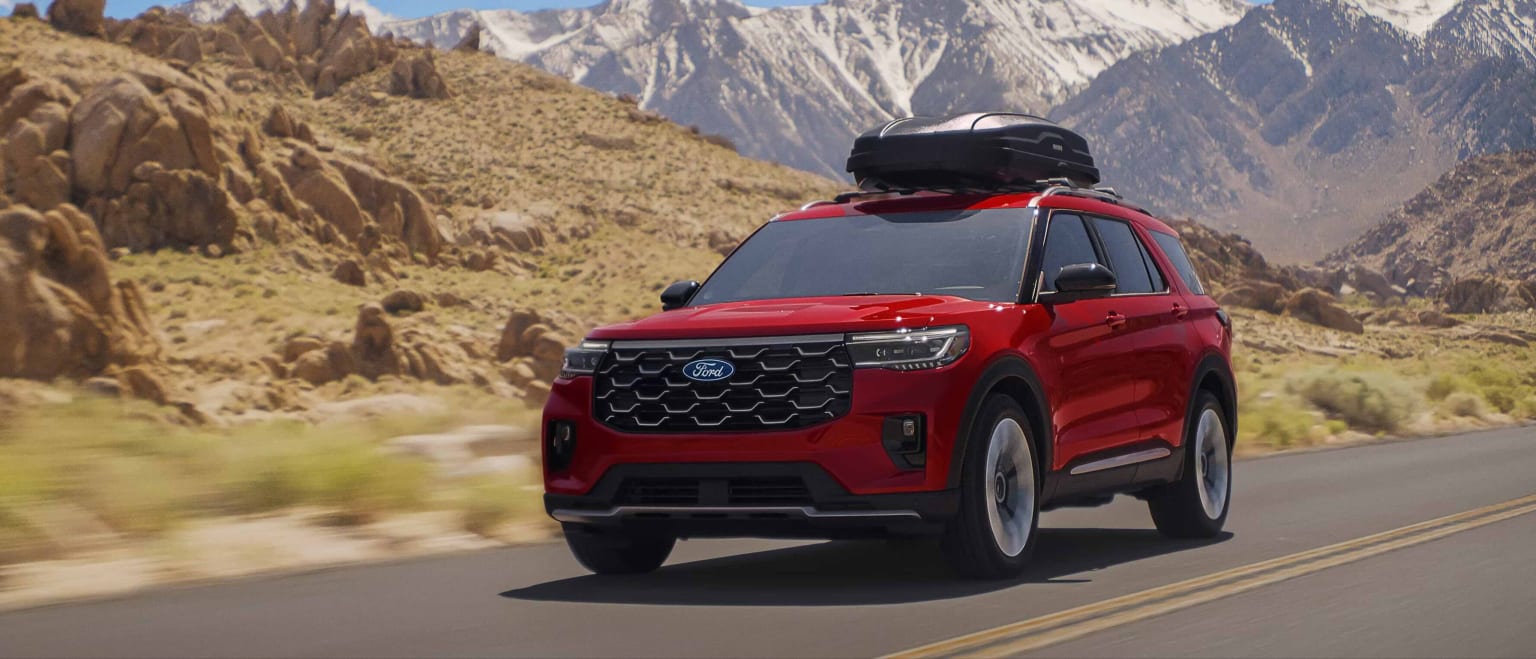 A red SUV with a roof-top carrier drives on a paved road through a desert landscape with snow-capped mountains in the background.