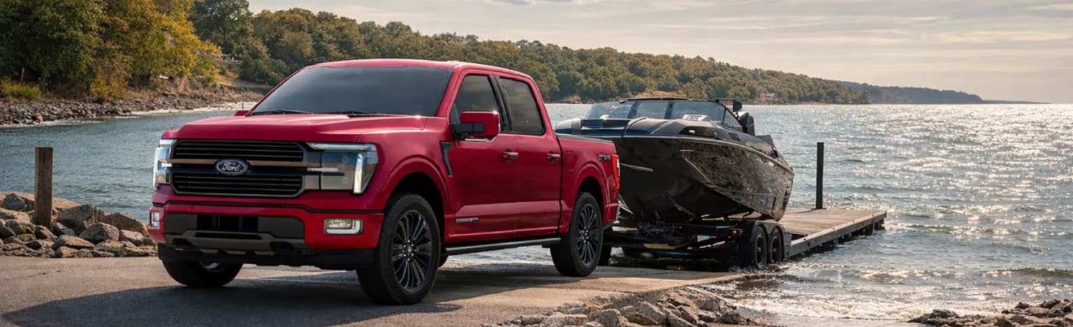 A red pickup truck towing a boat on a trailer down a ramp towards a lake, with a tree-covered shoreline in the background.