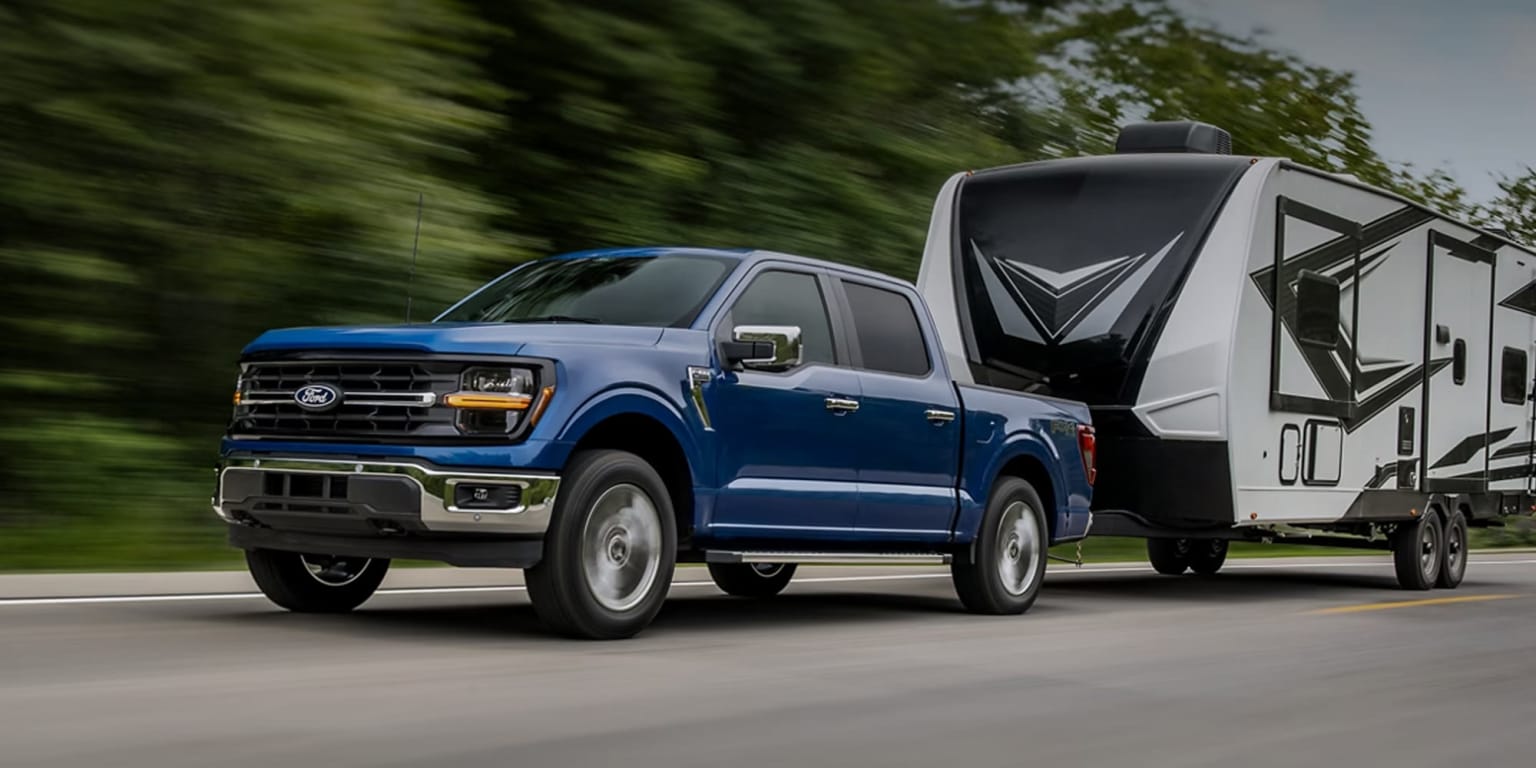 A blue pickup truck towing a large recreational vehicle on a blurred road surrounded by lush greenery.