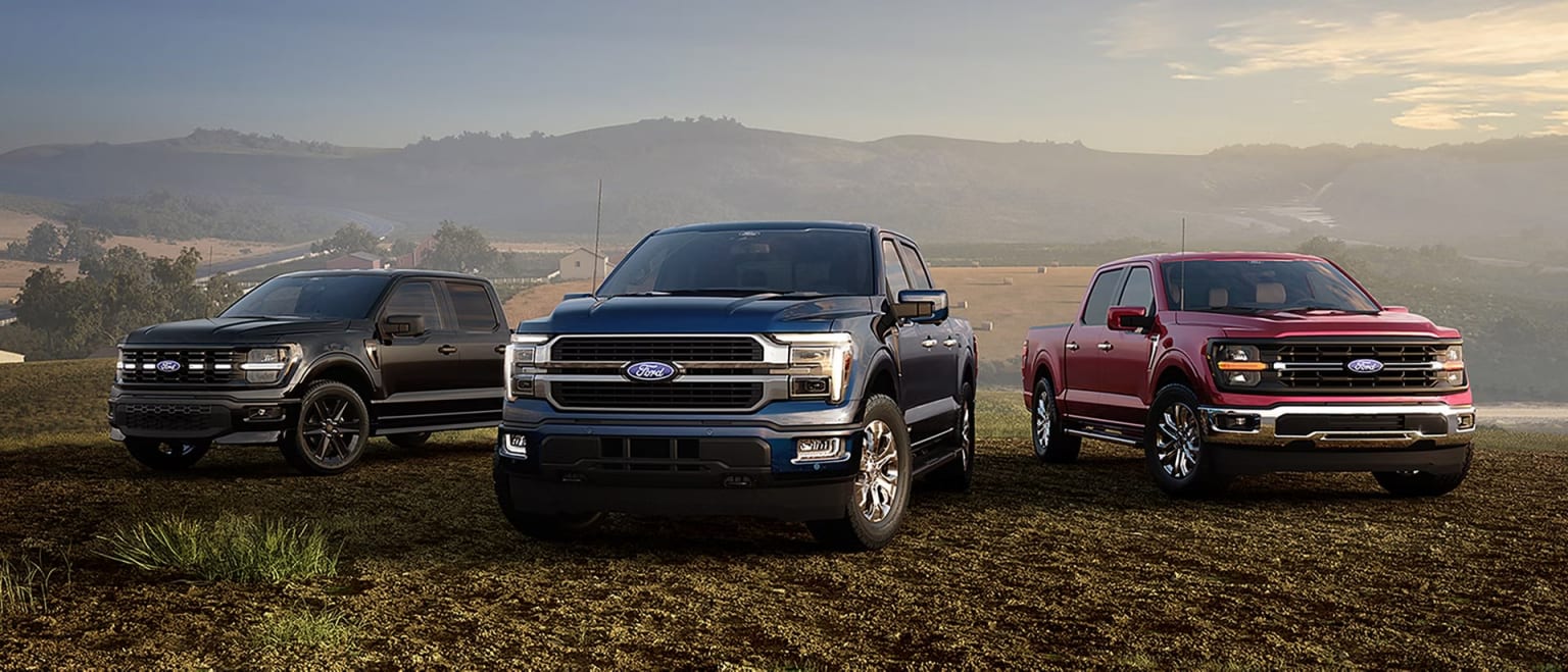 Three trucks are parked on a grassy field with rolling hills in the background.