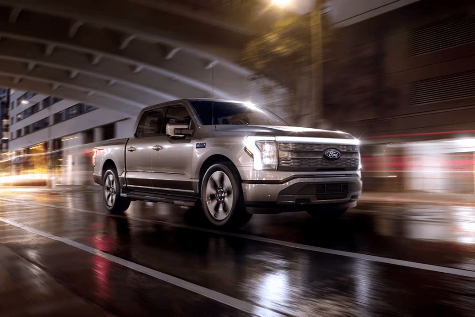 A silver pickup truck driving on a wet road at night with blurry lights and buildings in the background.
