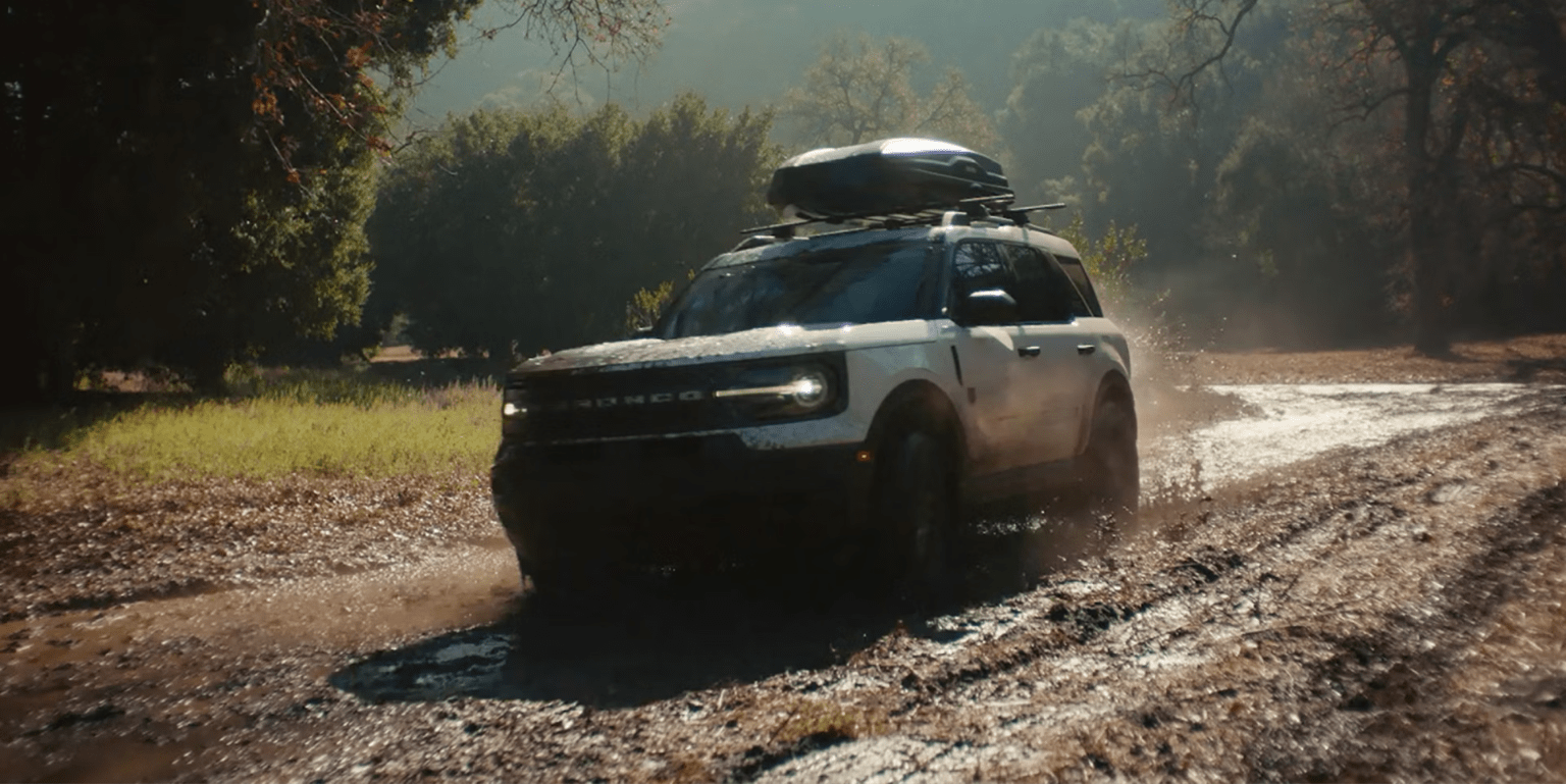 A white SUV with a roof rack drives through a muddy trail with trees in the background.