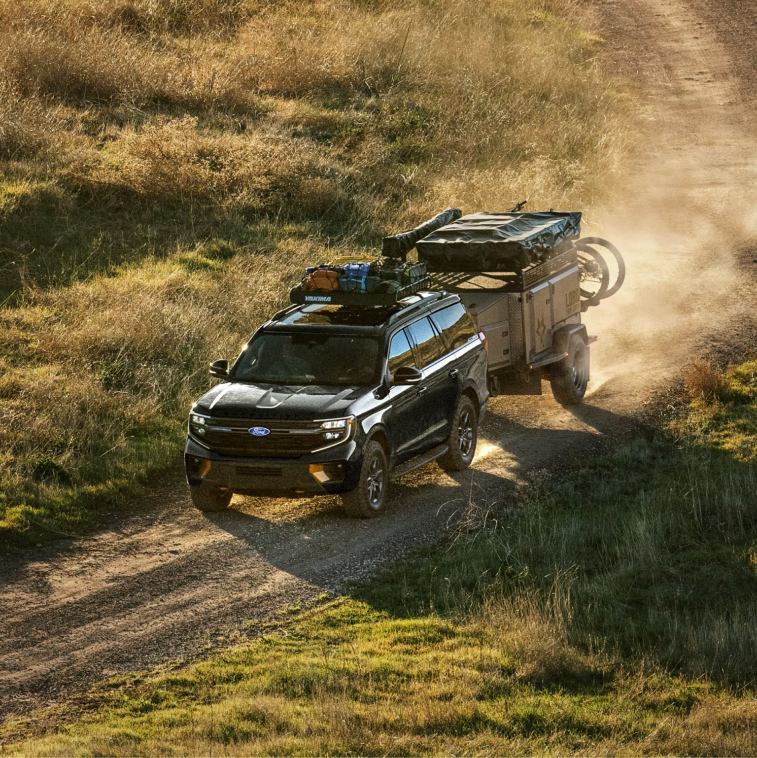 A black SUV towing a camping trailer kicks up dust while driving along a dirt path through a grassy field.