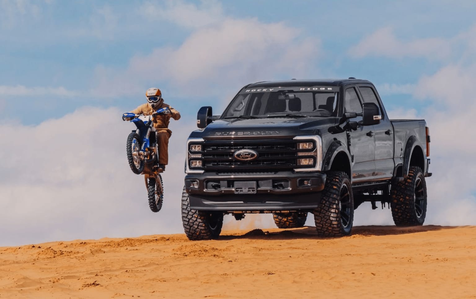 Off-road pickup truck with large tires and a person riding a motorcycle in the foreground, against a backdrop of a cloudy sky and sandy terrain.