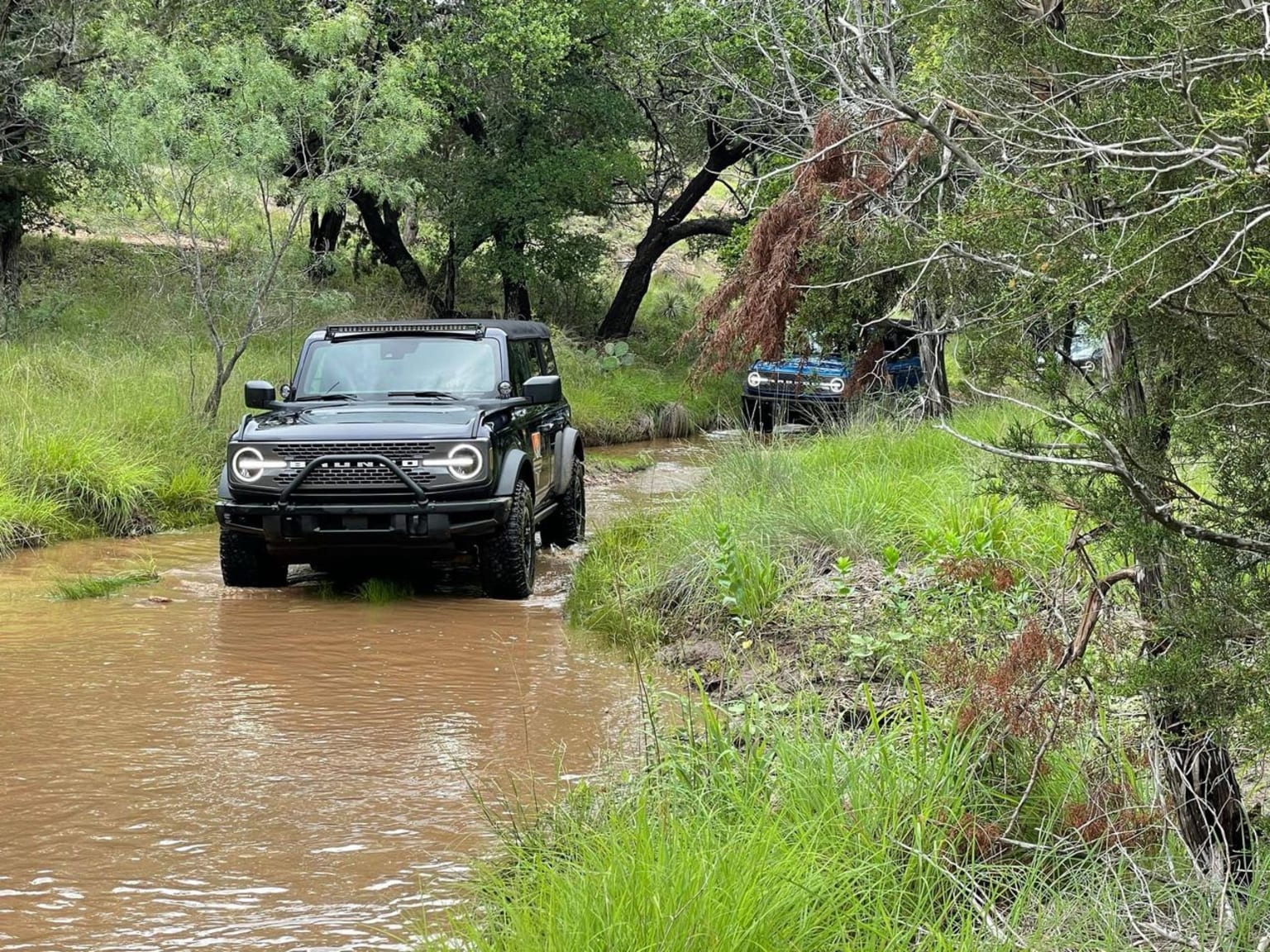 a jeep driving through a muddy river in the woods with a trailer in the back of the truck on the other side of the stream