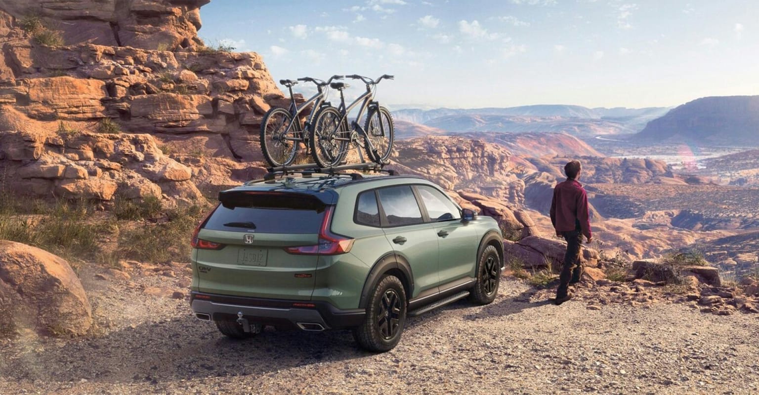 A green SUV with bikes on its roof is parked on a rocky trail, with a person standing beside it and a vast, arid landscape stretching into the distance.