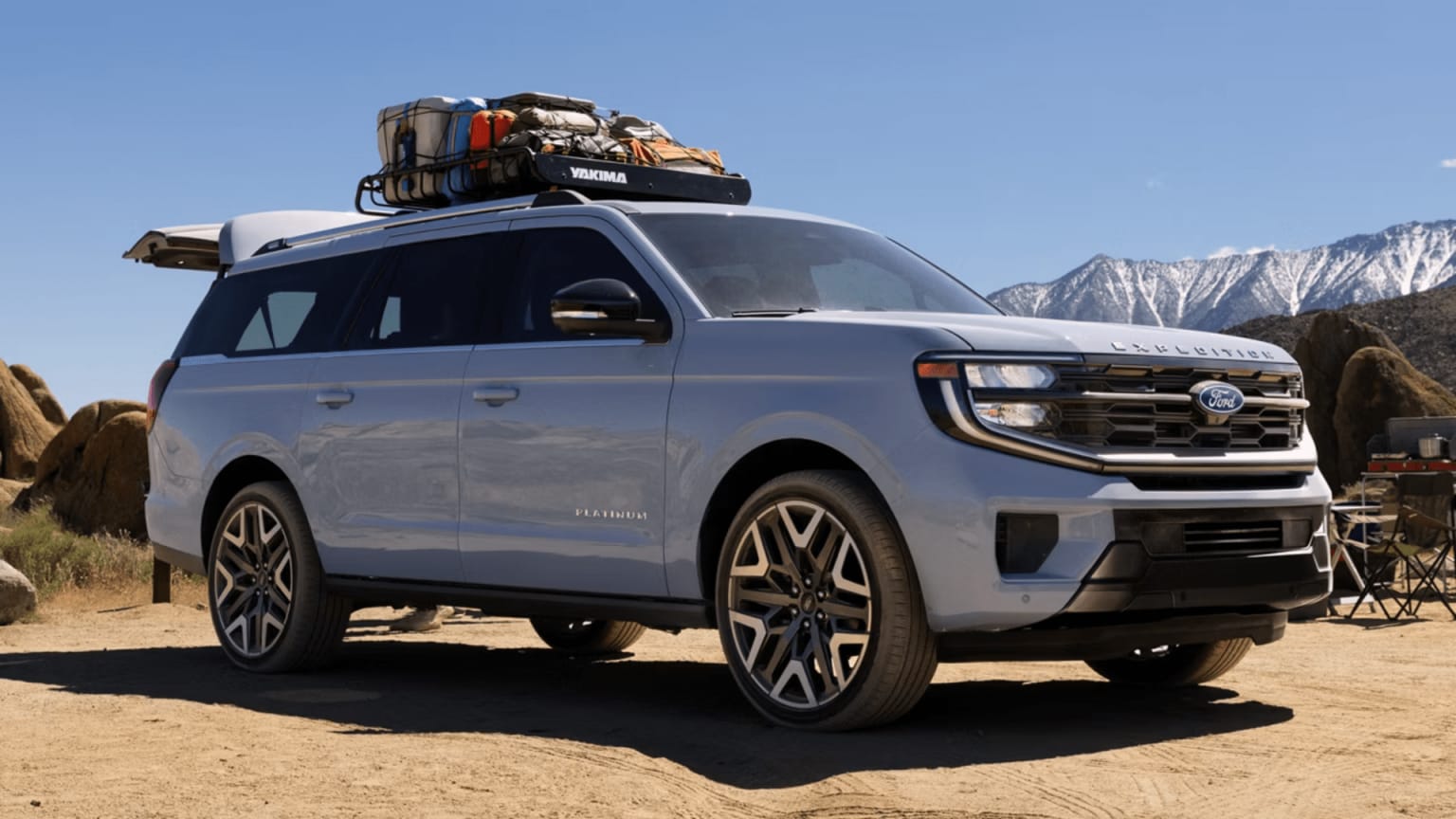 A light blue large SUV loaded with gear on its roof is parked on sandy ground against a backdrop of rocky terrain and snow-capped mountains.