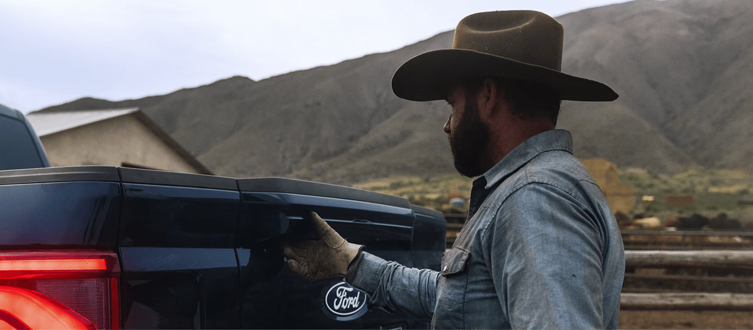 A cowboy opening the tailgate of a truck with a ranch and mountains in the background.