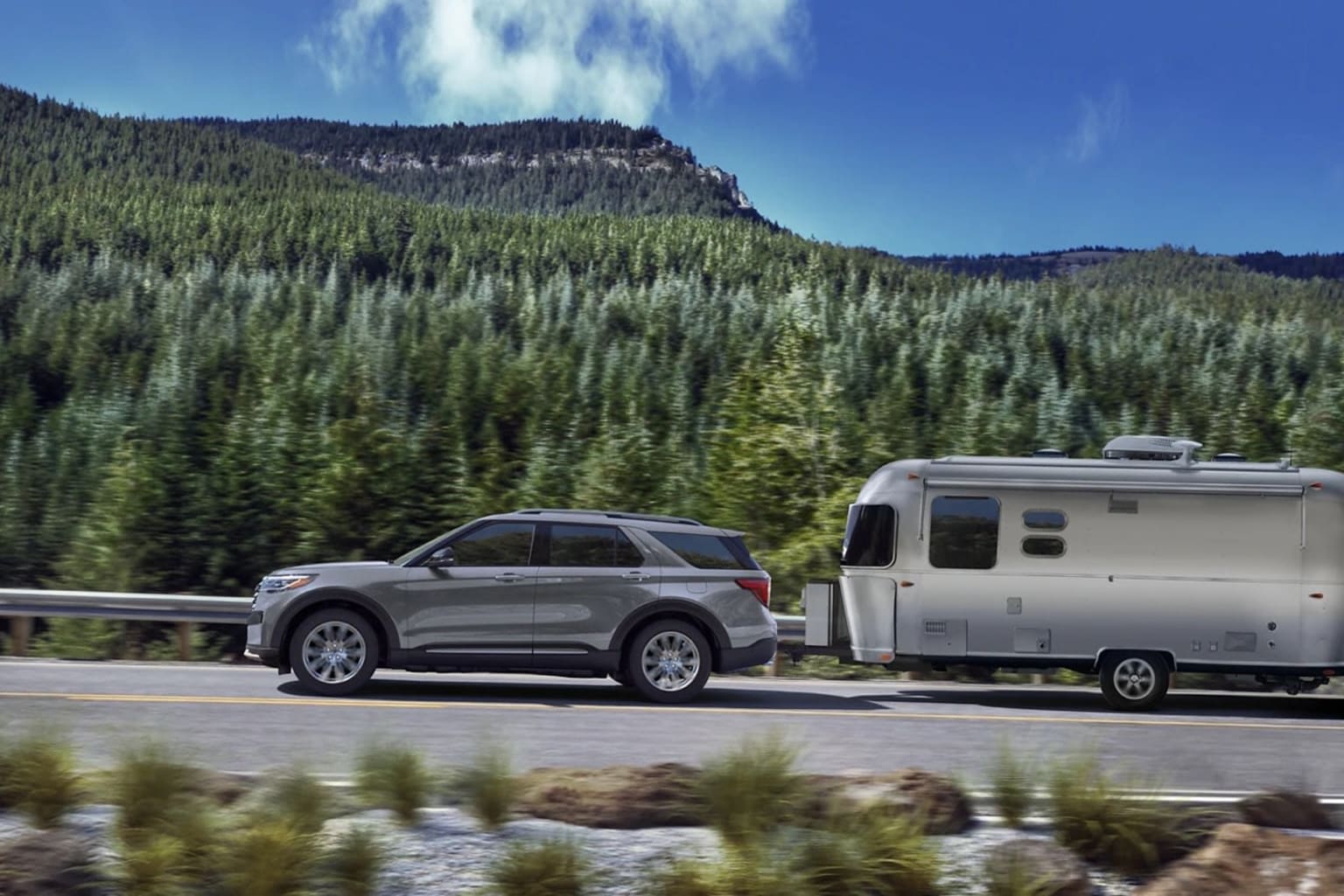 Large SUV towing a silver Airstream trailer traveling along a winding road through a forested mountain landscape with a rocky cliff in the background.
