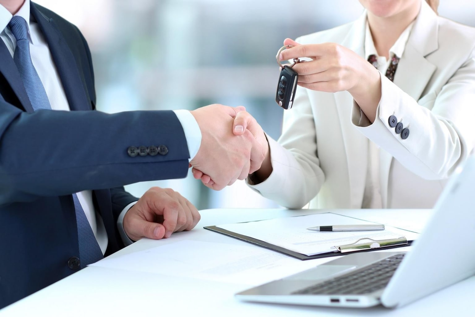 Two business professionals shaking hands over a desk in an office setting with a laptop and various office supplies visible in the background.