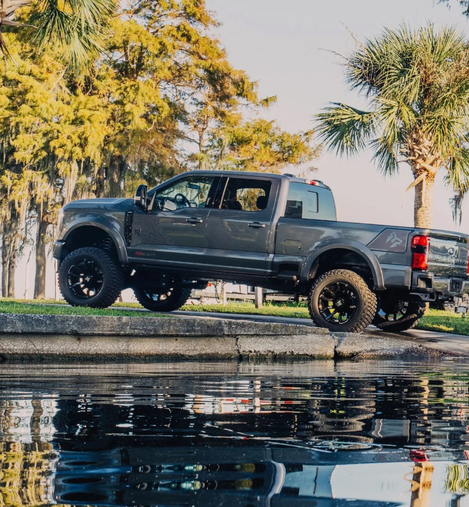 A large, lifted pickup truck is parked on a concrete ledge, its reflection visible in the still water below, surrounded by lush greenery and palm trees.