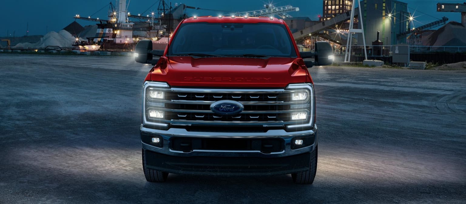 A large, red pickup truck with a rugged and powerful appearance is prominently featured in the foreground. The background shows a nighttime industrial setting with various structures and machinery visible in the distance.