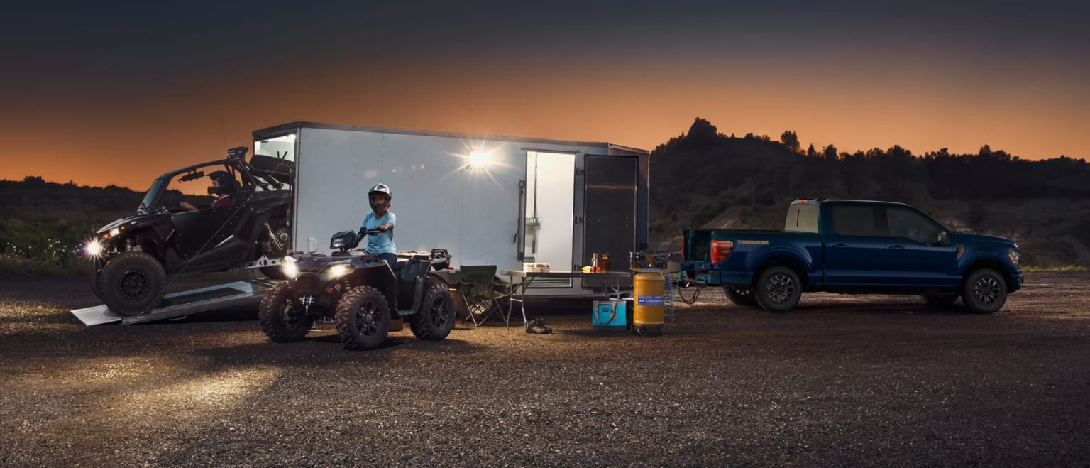 A nighttime outdoor scene with a large truck, a trailer, and a person standing near the trailer, set against a backdrop of a mountainous landscape and a colorful sunset sky.