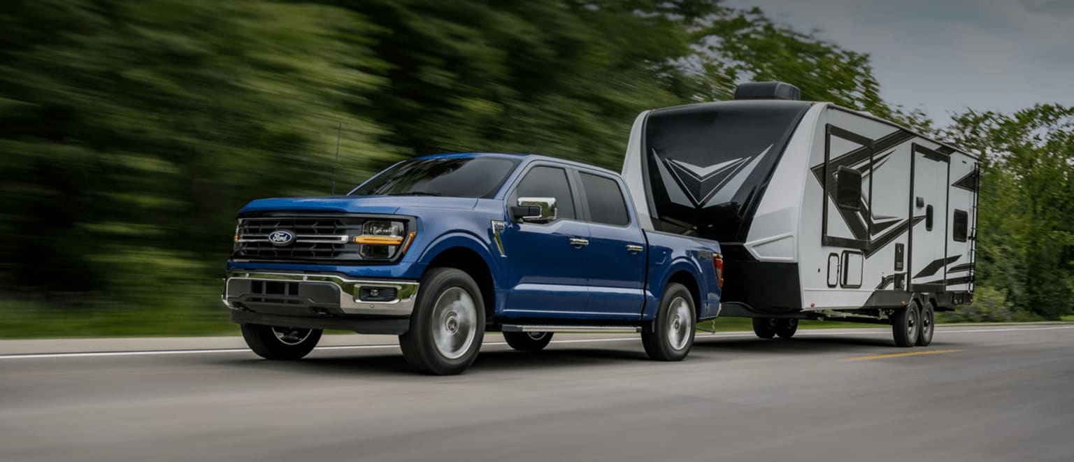 A blue pickup truck is towing a large recreational vehicle on a winding road surrounded by lush green foliage.