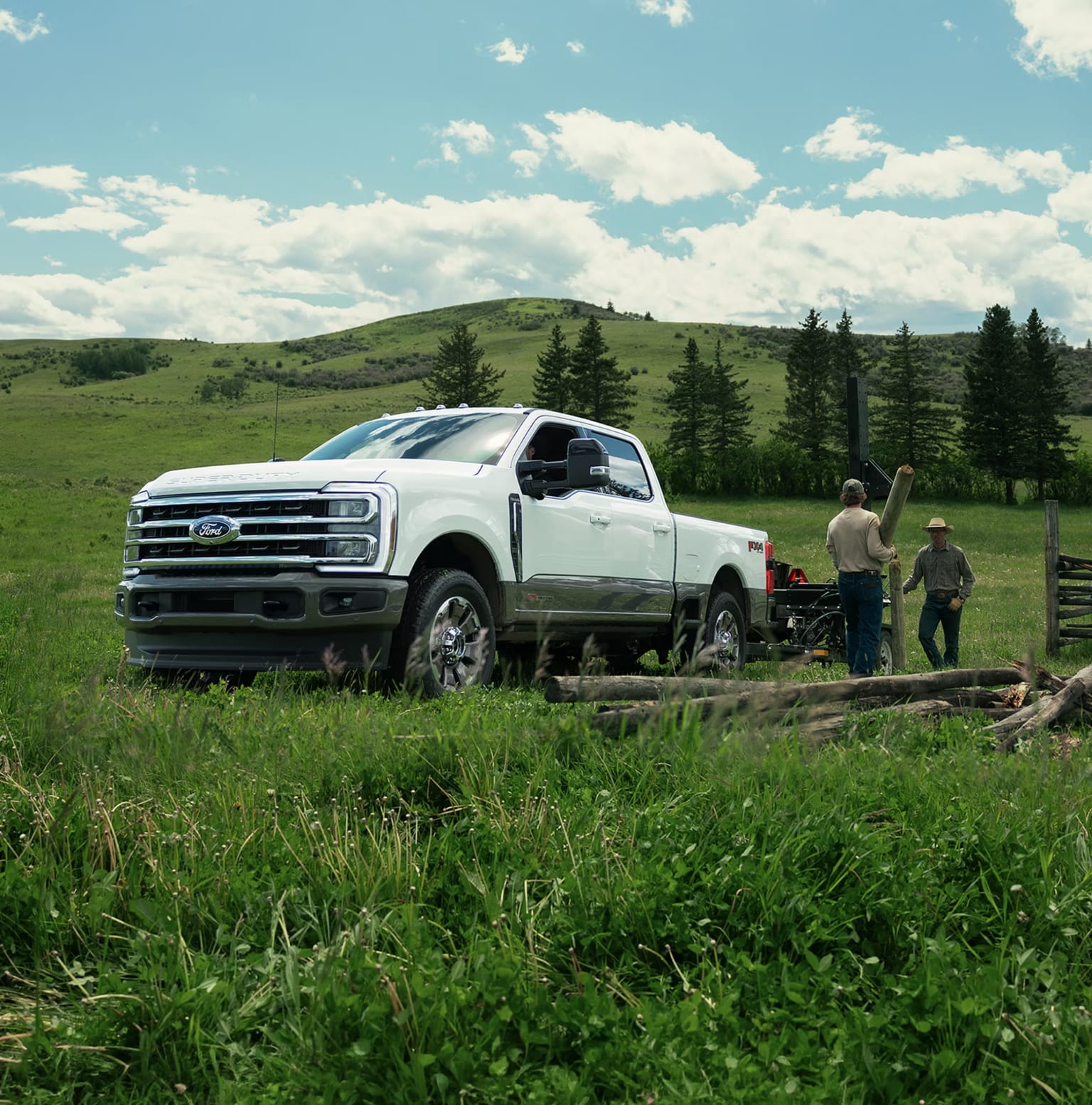 A white pickup truck in a grassy field with two men nearby, against a backdrop of green hills and cloudy sky.