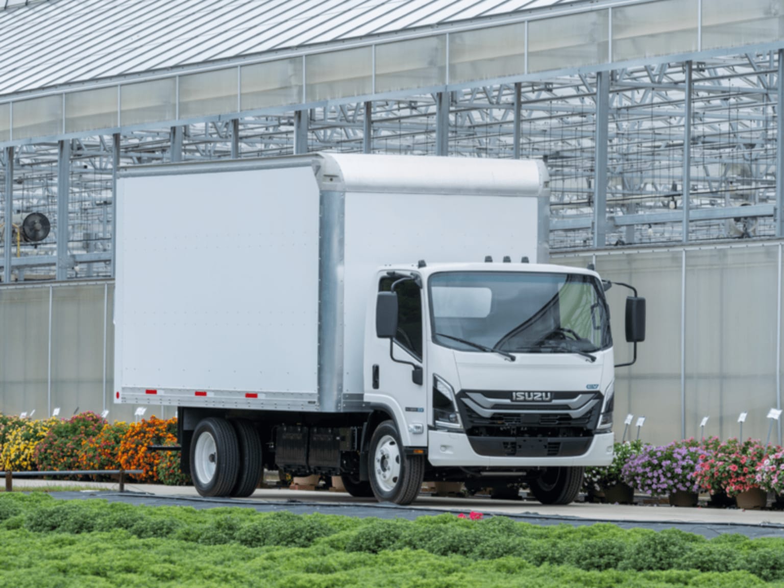 A white box truck parked on a paved path next to green foliage and colorful flowers, with a large glass greenhouse in the background.