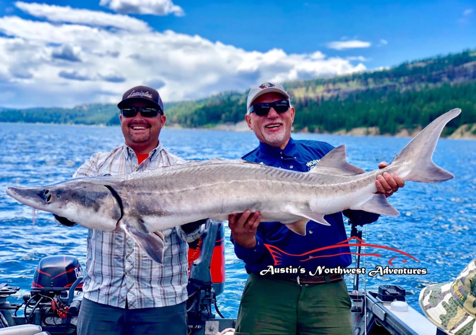 Two men in a boat with a large fish in the foreground, serene lake, hills, and cloudy sky in the background.
