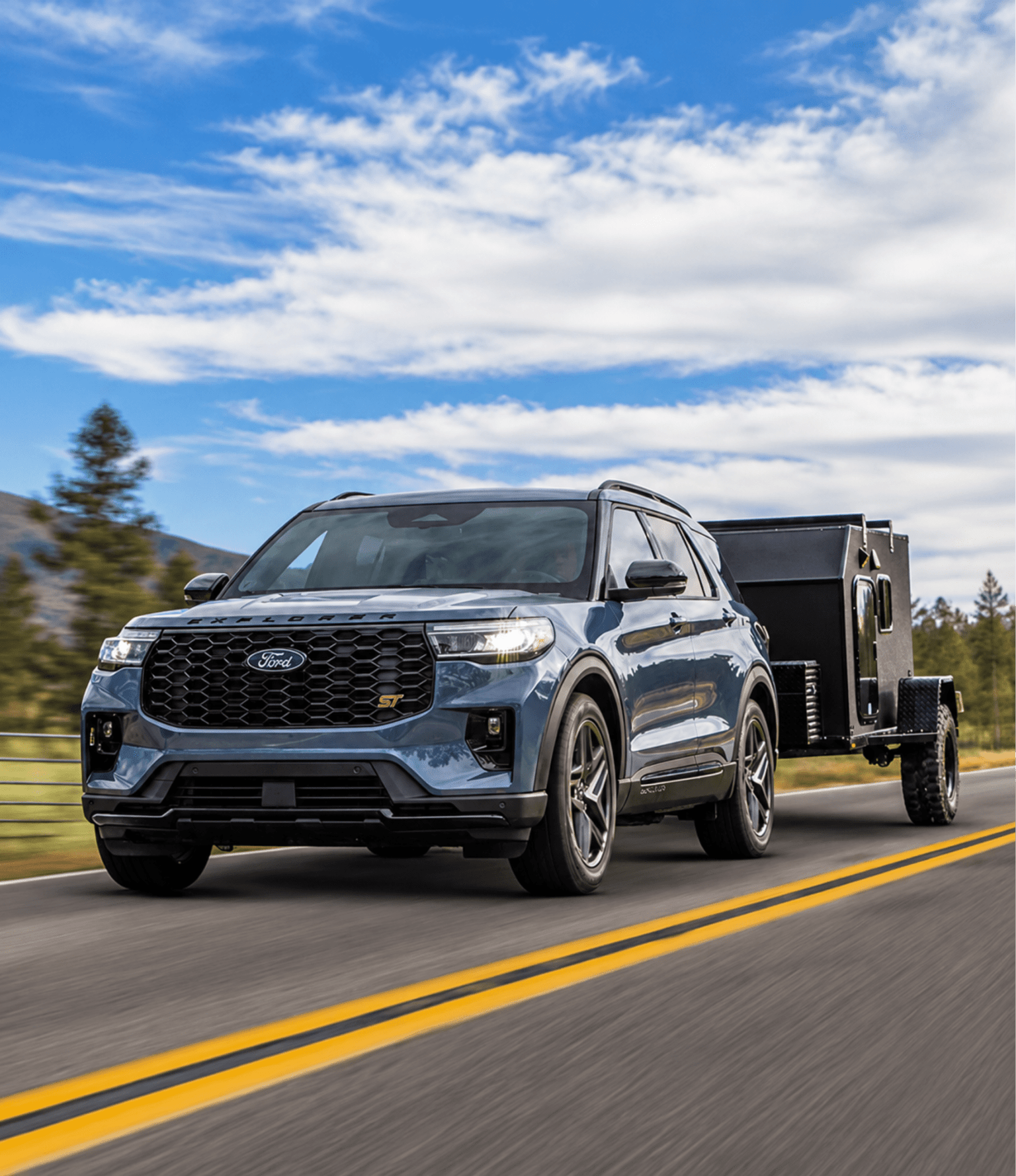 A blue SUV pulls a black utility trailer along a paved road under a bright, cloudy sky.