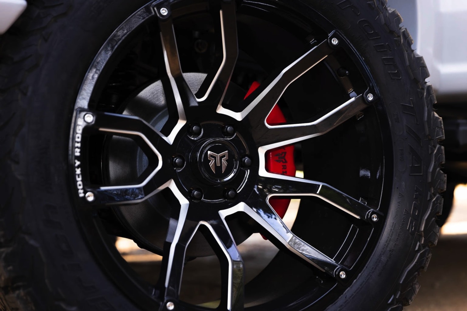 A close-up view of a black alloy wheel with a distinctive design, featuring red brake calipers visible behind the spokes. The background is out of focus, emphasizing the wheel.