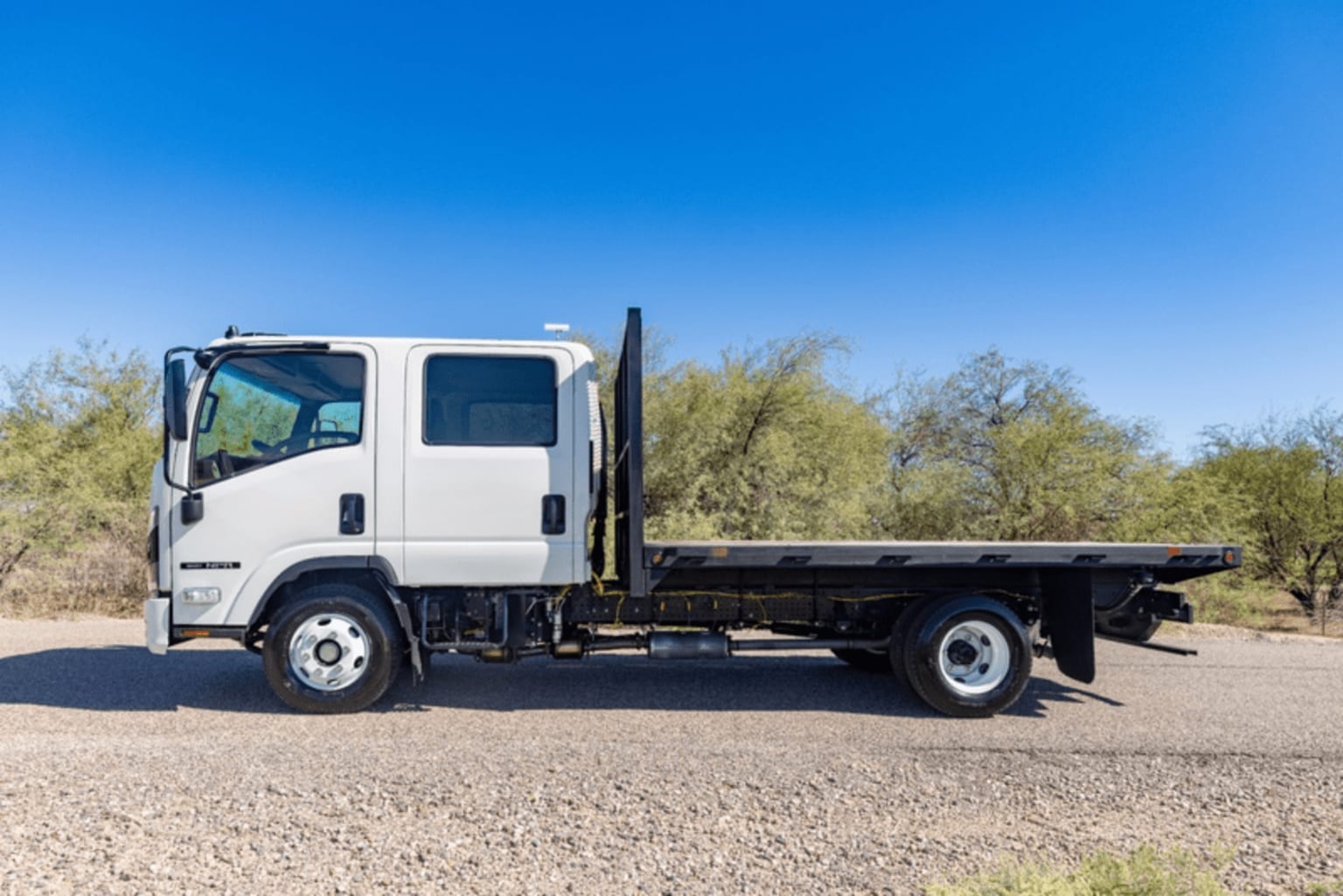 A white flatbed truck parked on gravel surface with green shrubs and clear blue sky in the background.