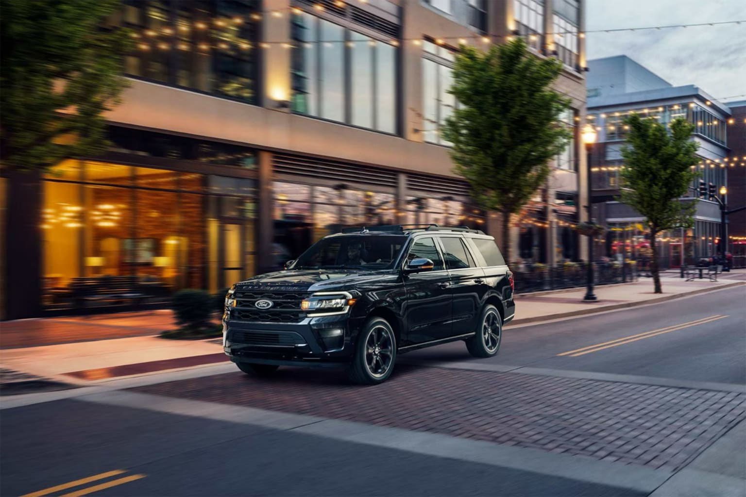 A large black SUV driving down a city street with buildings, trees, and street lamps in the background.