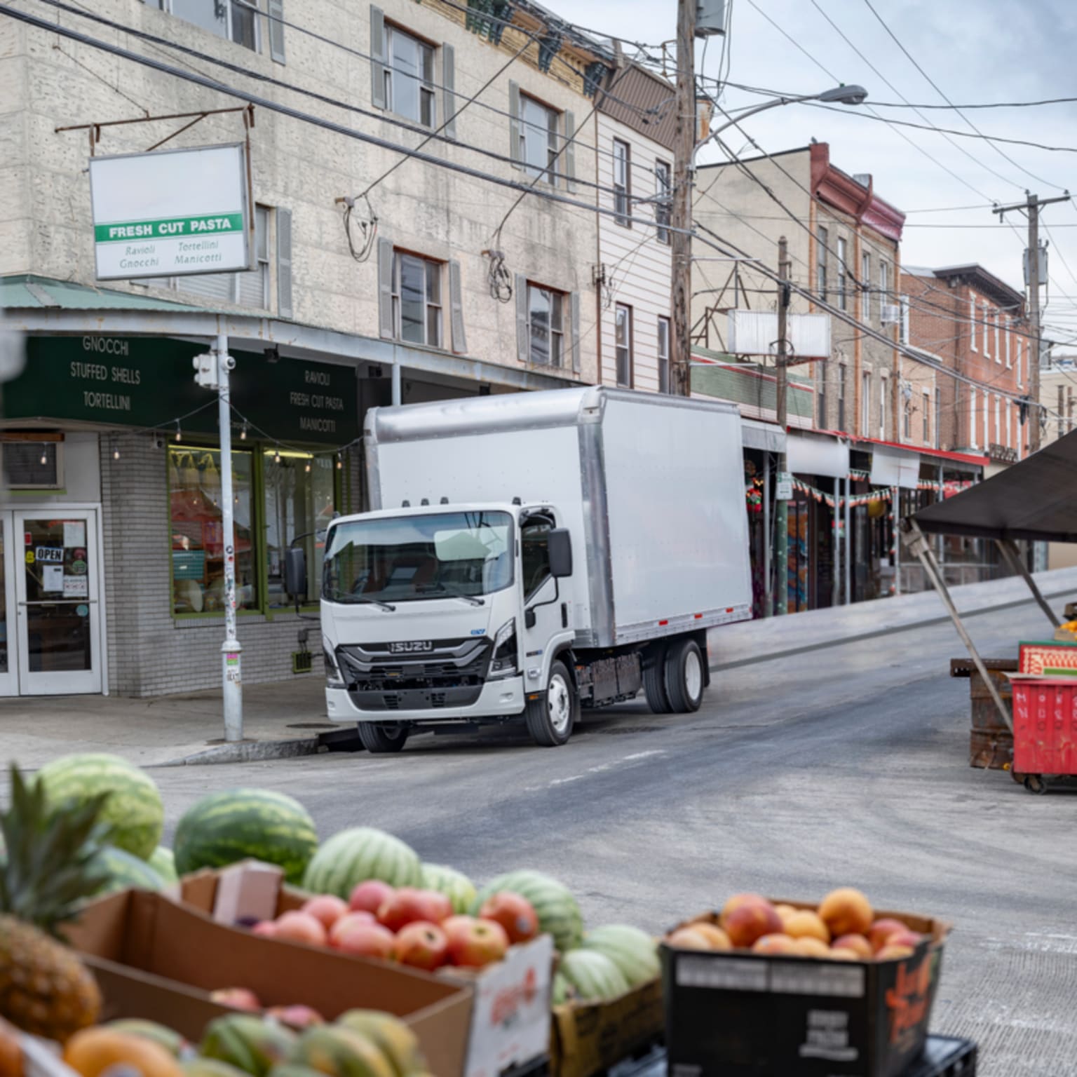 Fresh produce in crates fills the foreground, while a white box truck is parked on a city street lined with storefronts in the background.