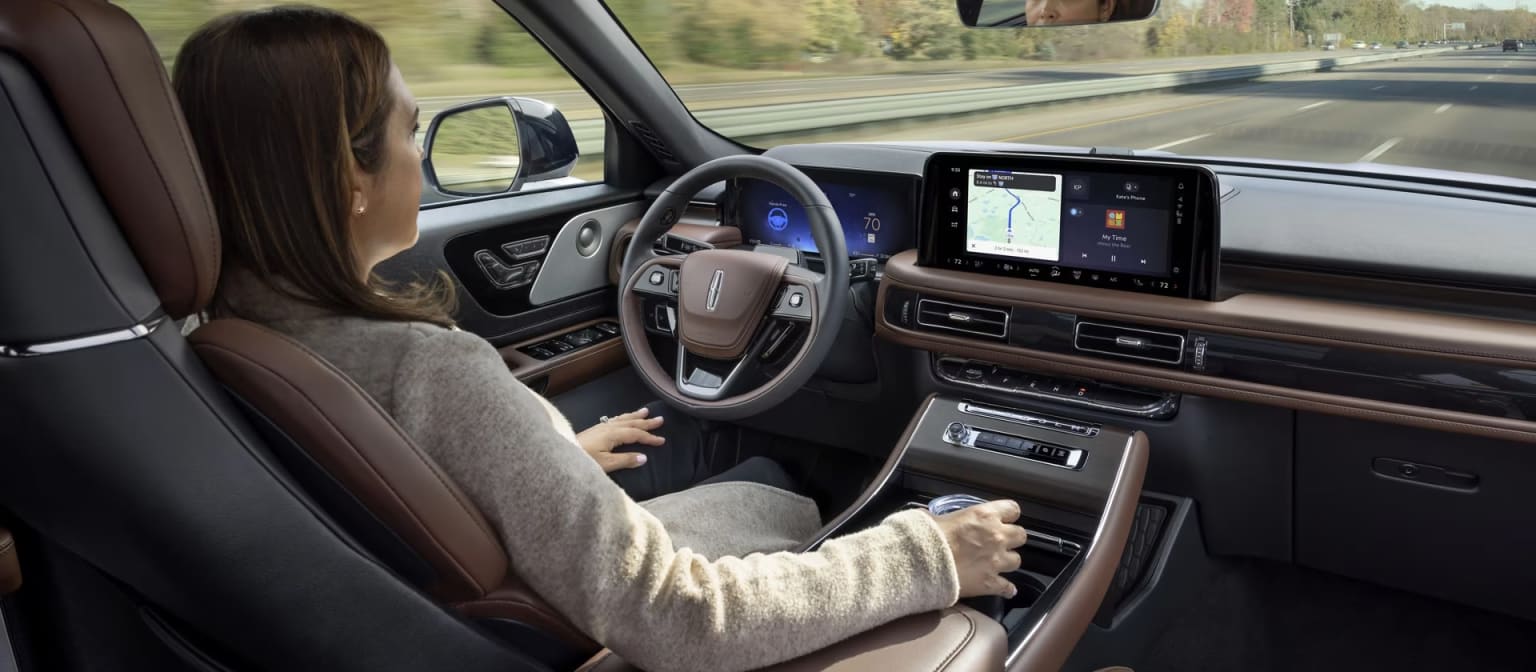 The image shows the interior of a vehicle with a person sitting in the driver's seat, various controls, and displays including a large touchscreen display. The background outside the vehicle appears to be a rural or forested area.