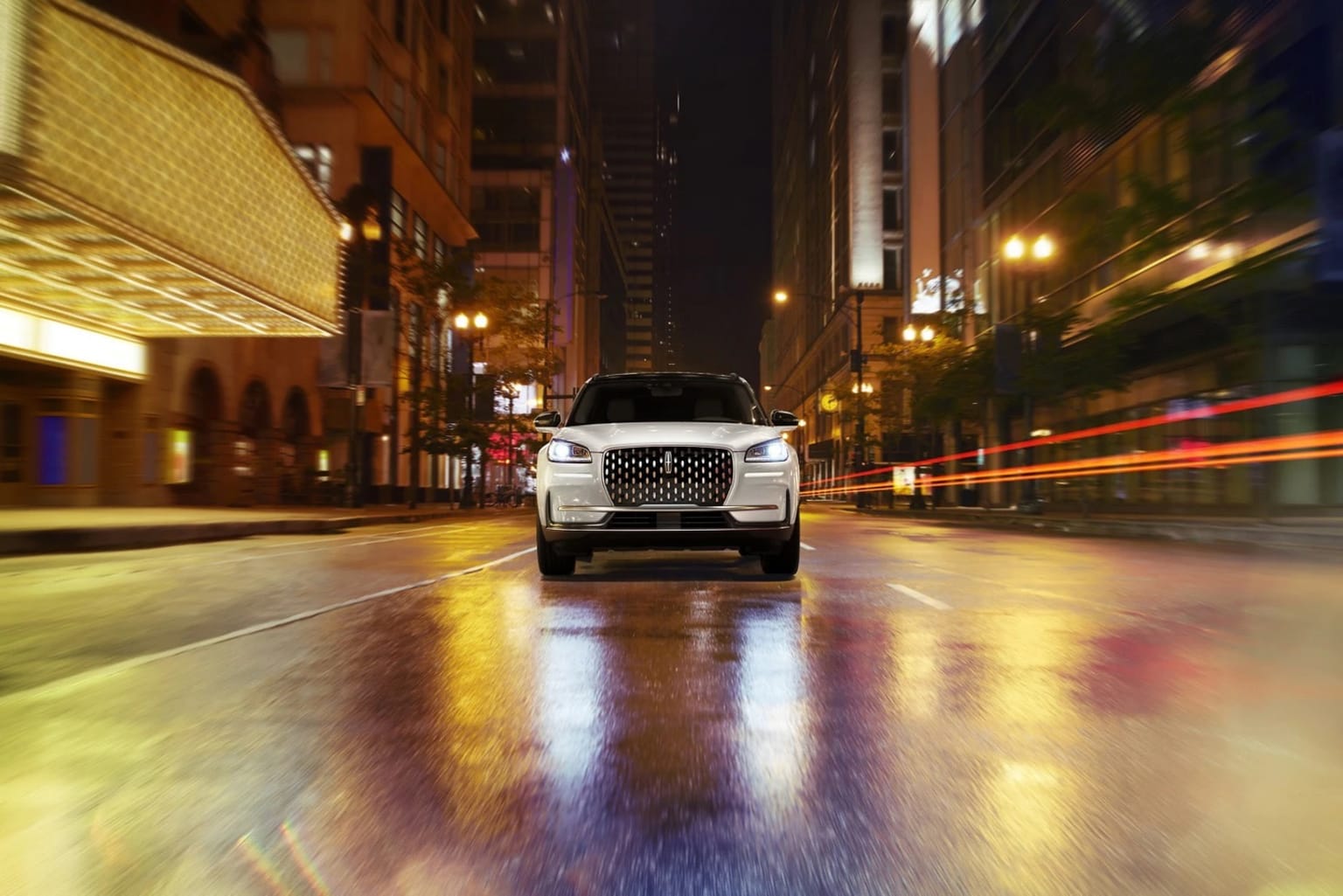 Front profile view of a white 2026 Lincoln Corsair driving down a big city street lit up by neon lights at night