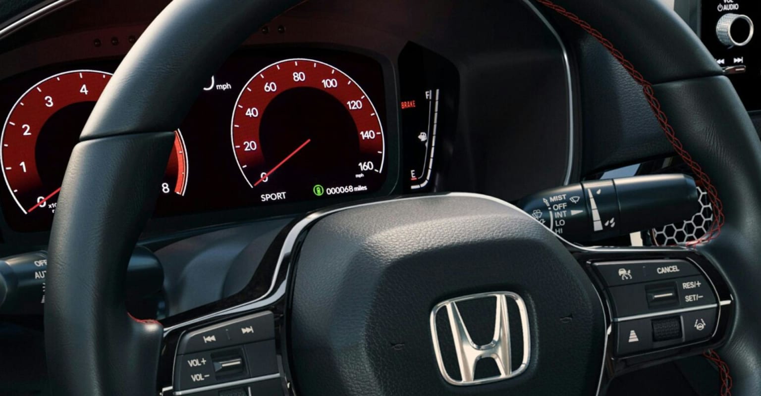 Close-up of a car's steering wheel and dashboard with speedometer and tachometer in the foreground, and blurred interior in the background.