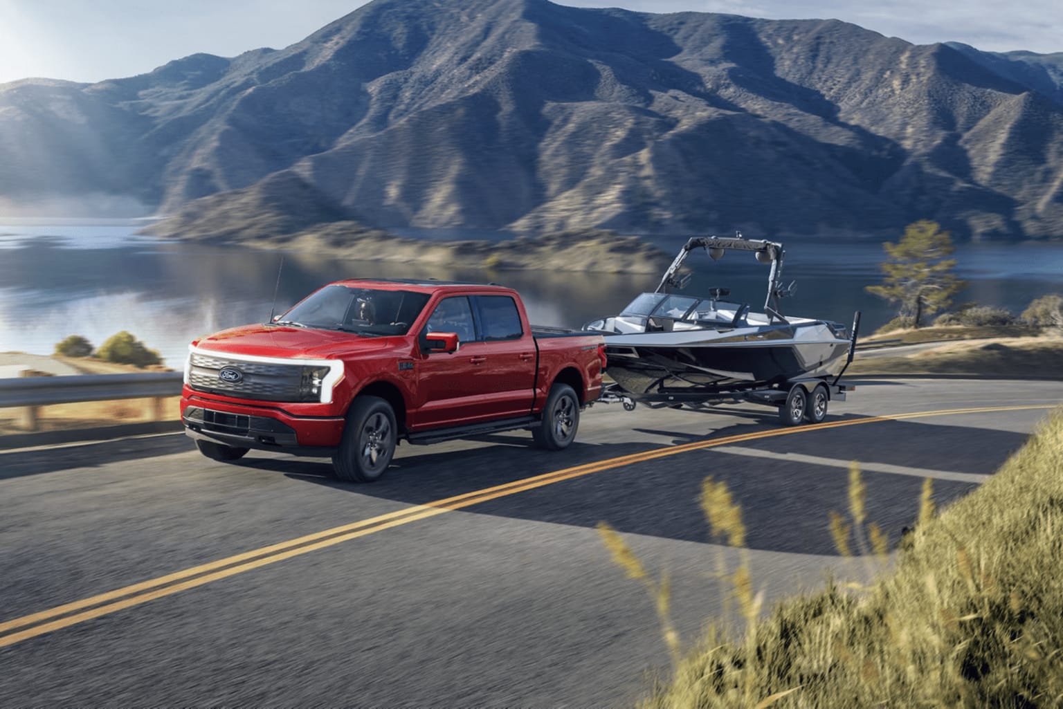 A red pickup truck towing a boat on a trailer in a mountainous landscape with a lake in the distance.