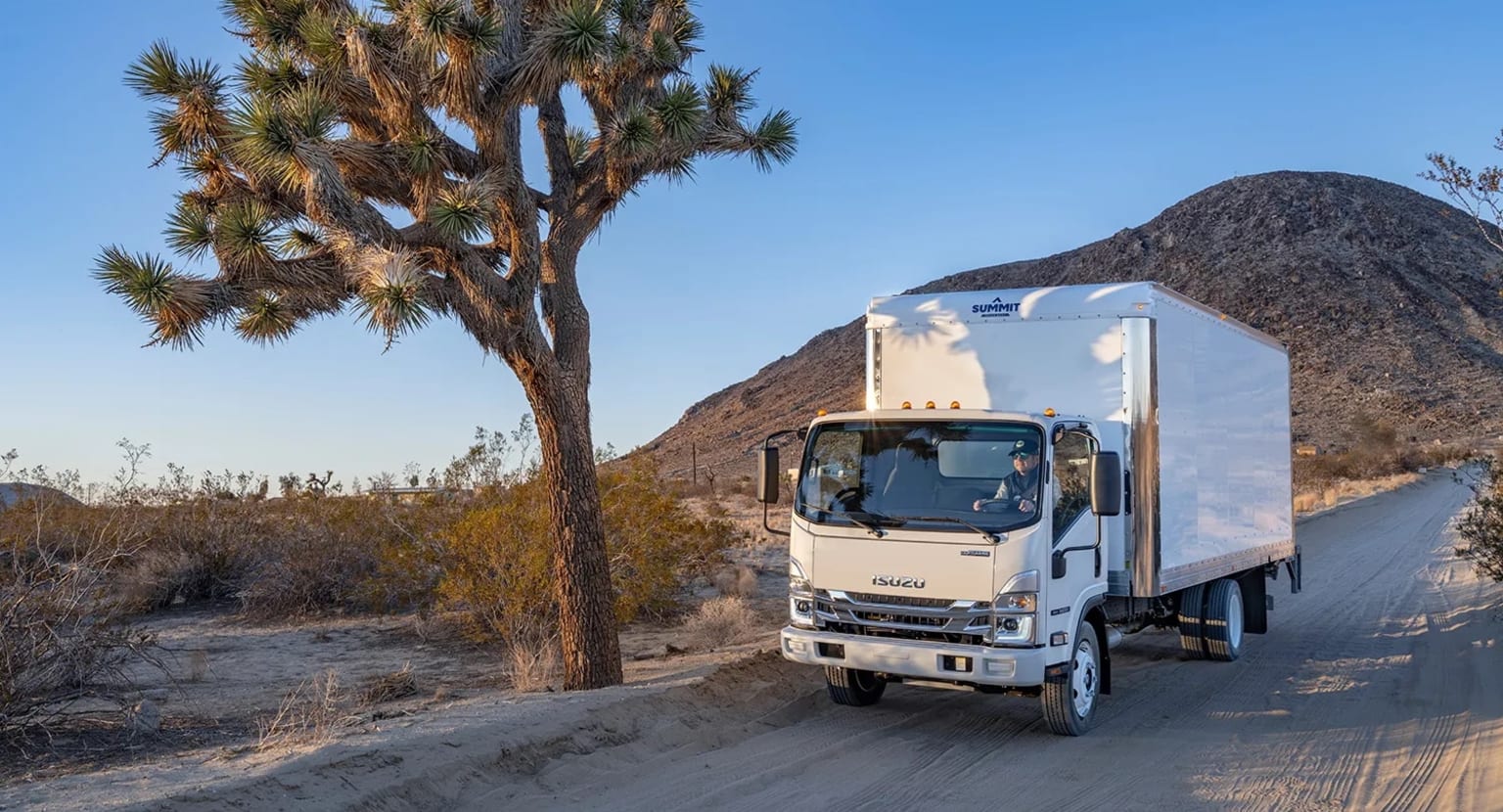 A large white delivery truck drives down a dirt road surrounded by Joshua trees and rugged mountains in the background.
