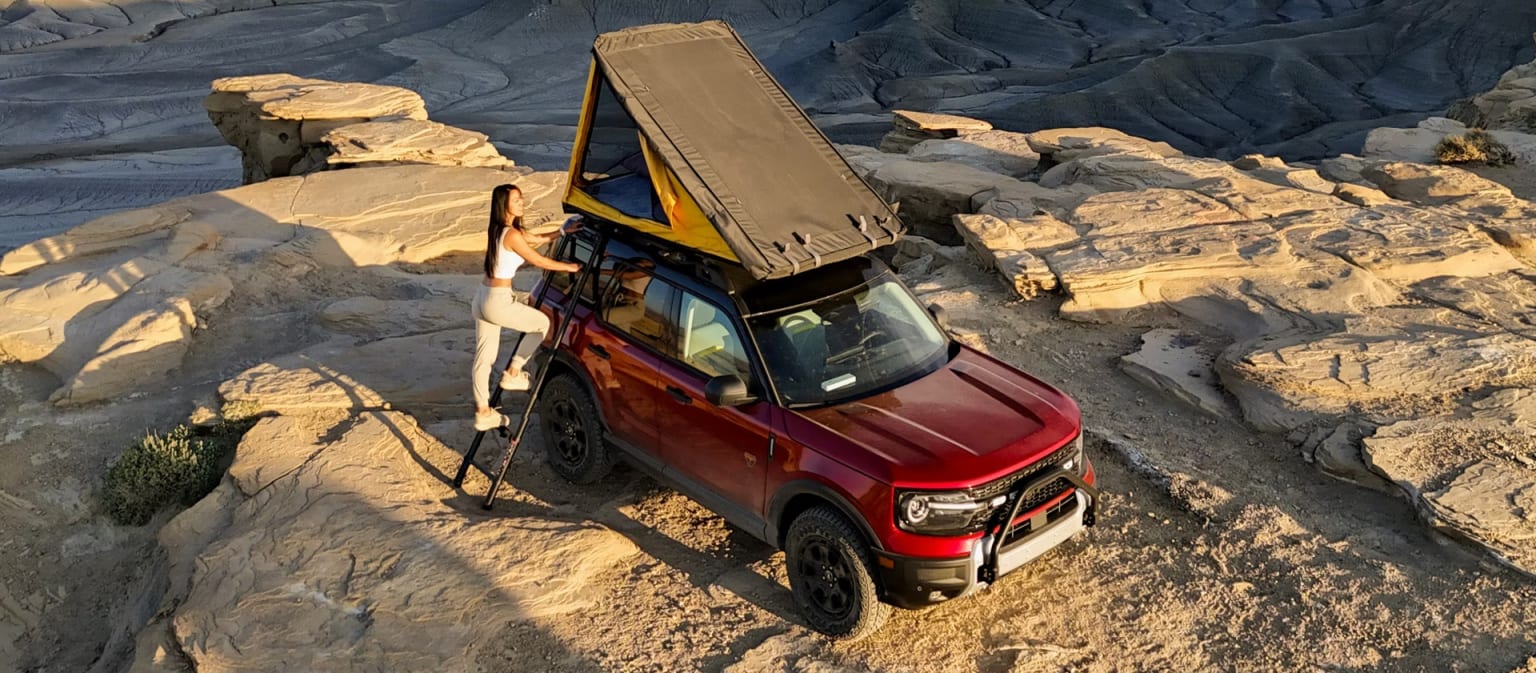 A red off-road vehicle with a large cargo box on its roof is navigating through a rugged, rocky terrain with a mountainous backdrop.