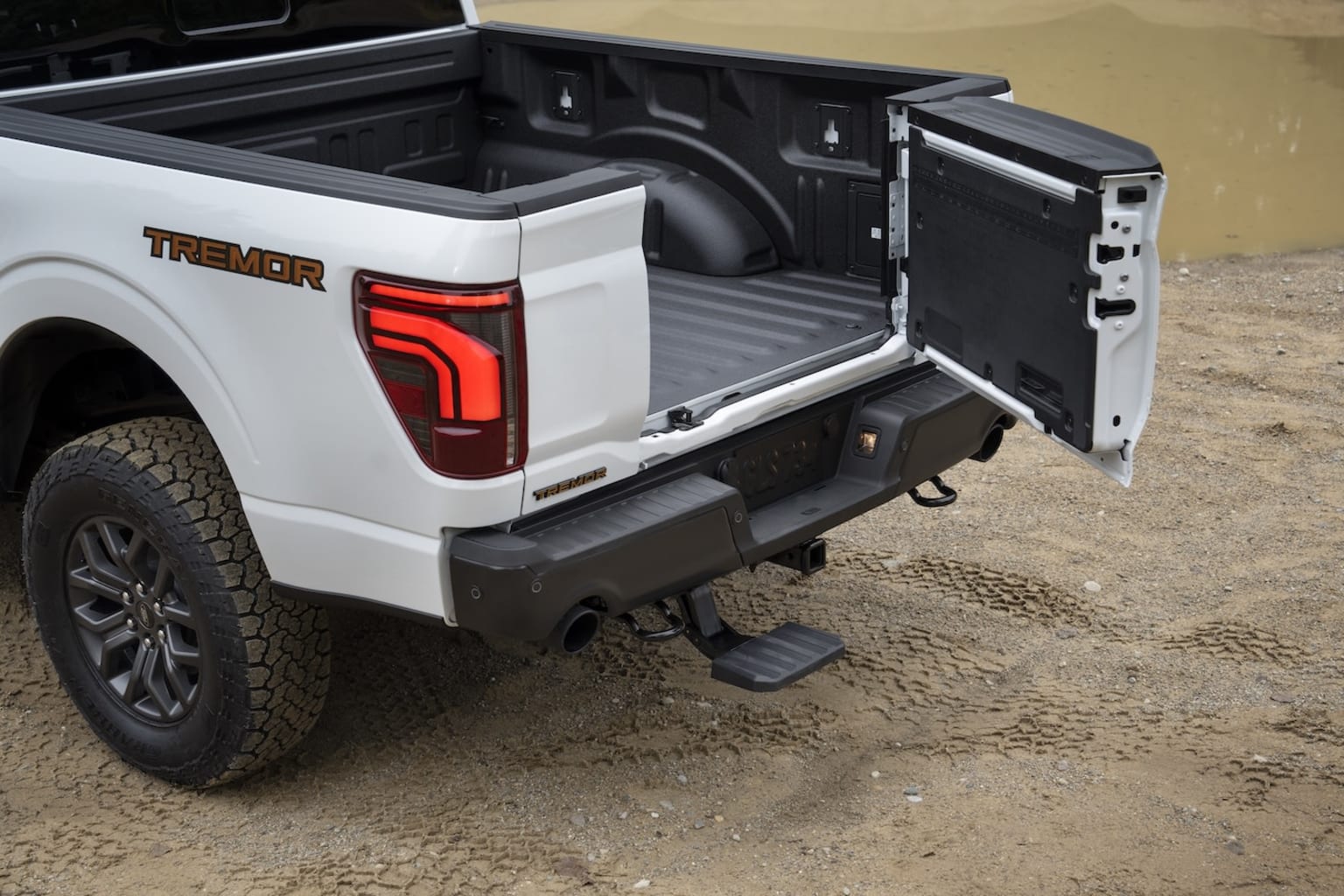 Rear view of a white pickup truck with the tailgate open, showcasing a roomy cargo bed. The truck is parked on a gravel or dirt surface, indicating an outdoor location.