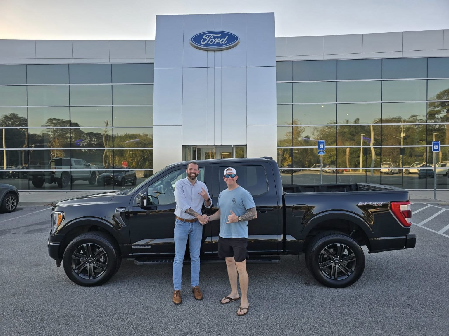 The image shows two people standing in front of a large dealership building, with a black pickup truck in the foreground.