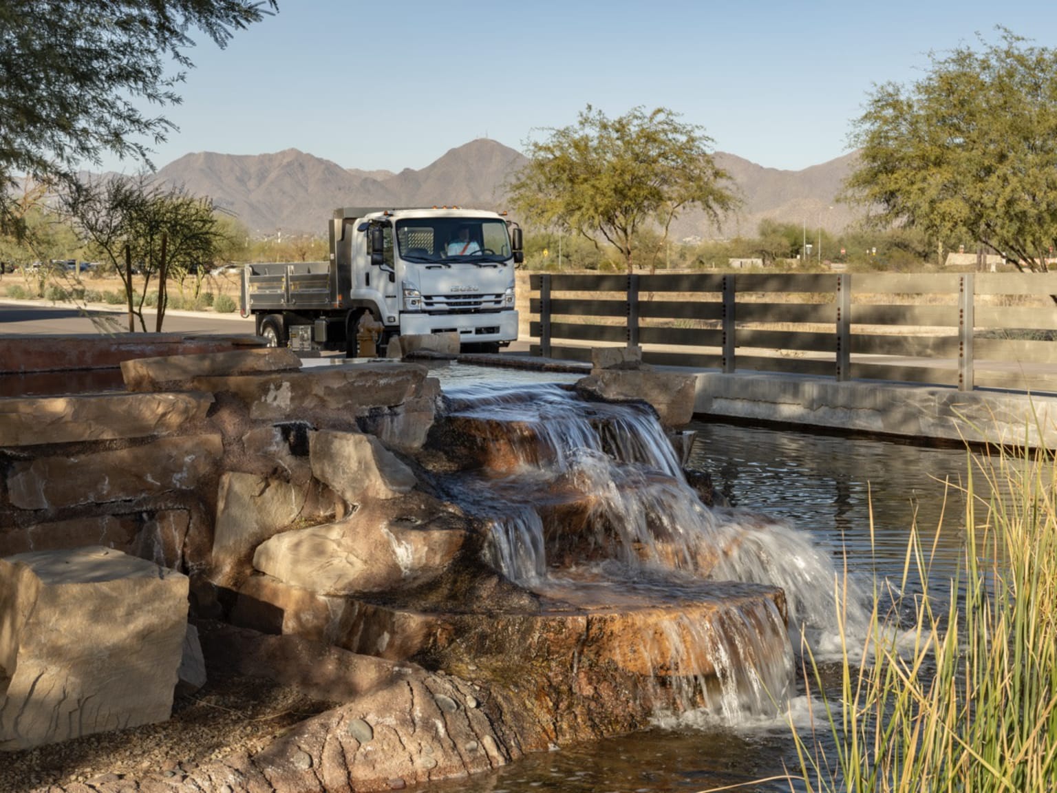A white dump truck parked near a man-made waterfall with rocky landscaping in the foreground and a wooden fence and mountains in the background.