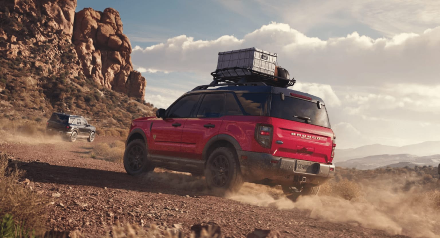 An off-road vehicle with a cargo box on the roof driving through a dusty, rocky landscape with towering cliffs in the background.