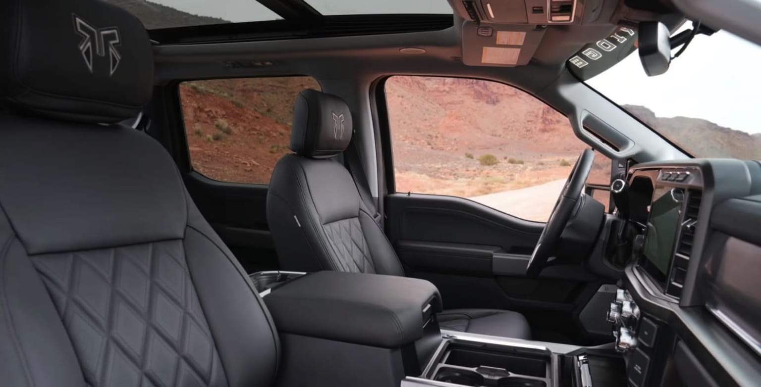 Interior of a vehicle with a dashboard, steering wheel, and seats. Background shows a mountainous or hilly landscape.