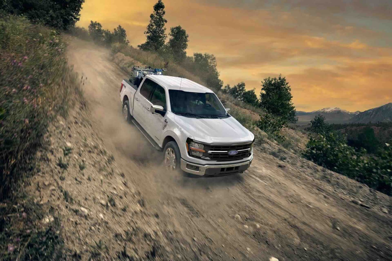 A white pickup truck driving down a steep, rocky dirt road with an hazy, orange sunset in the background.