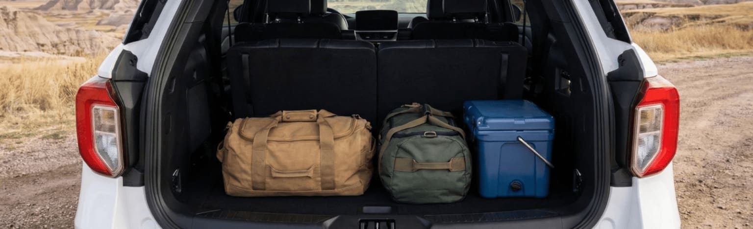 Three bags and a cooler are packed in the open trunk of a white SUV, with a dry, hilly landscape in the background.