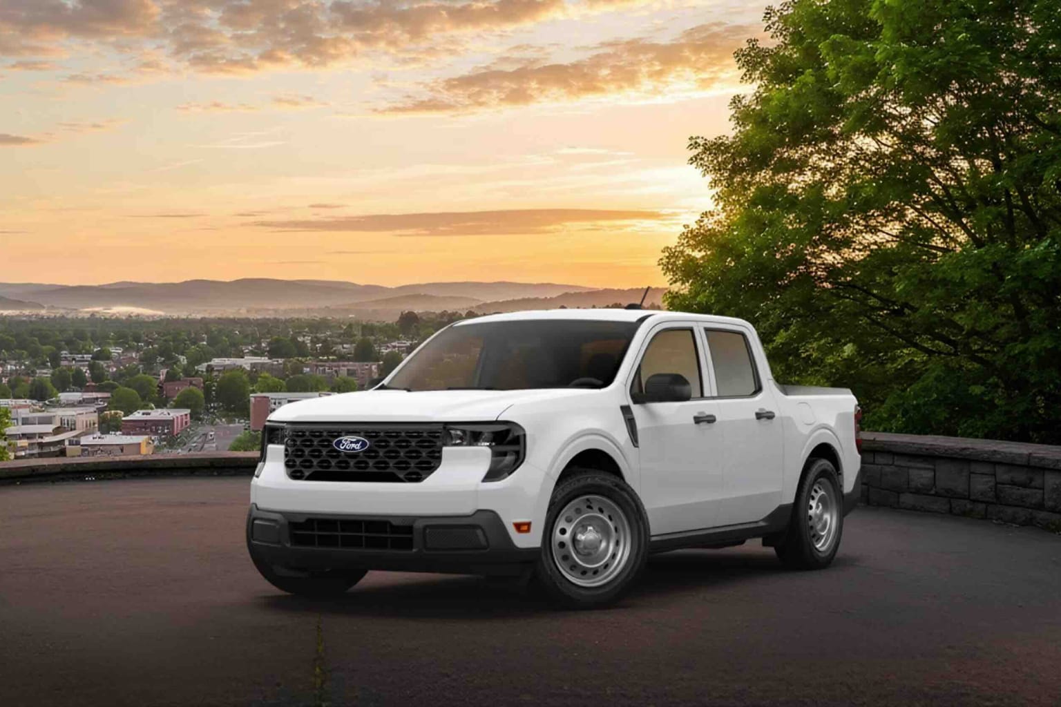 A white pickup truck on a paved surface with a scenic view of a town and rolling hills under a sunset sky in the background.