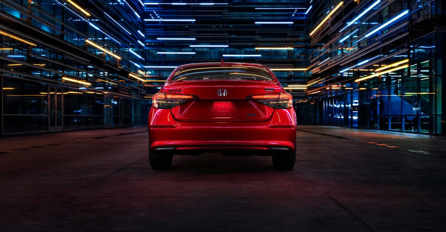 A red sedan is parked in a dark alleyway illuminated by colorful neon lights.