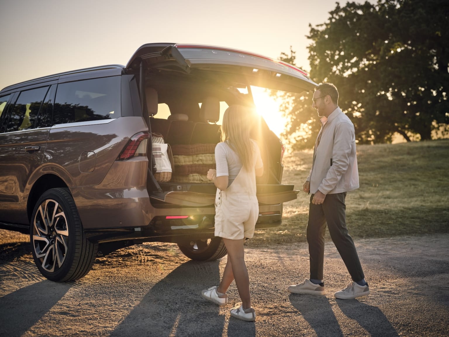 A couple standing next to a parked SUV in a grassy field, with the sun setting in the background, creating a warm, golden glow.