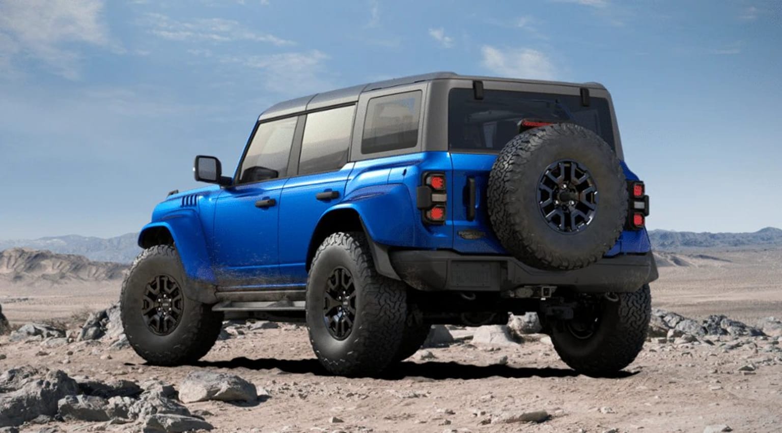 An off-road vehicle with large tires and a rugged exterior is parked on a rocky, desert-like terrain against a backdrop of a clear blue sky with scattered clouds.